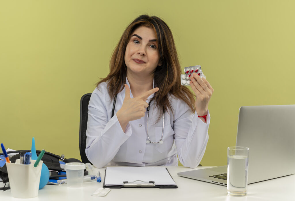 pleased middle-aged female doctor wearing medical robe with stethoscope sitting at desk work on laptop with nedical tools holding and points to pills on isolated green background with copy space