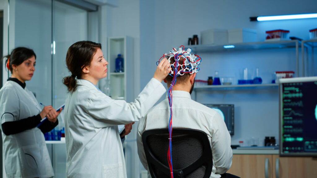 Brain Aneurysm: Back view of man patient wearing performant brainwave scanning headset sitting in neurological research laboratory while medical researcher adjusting it, examining nervous system 